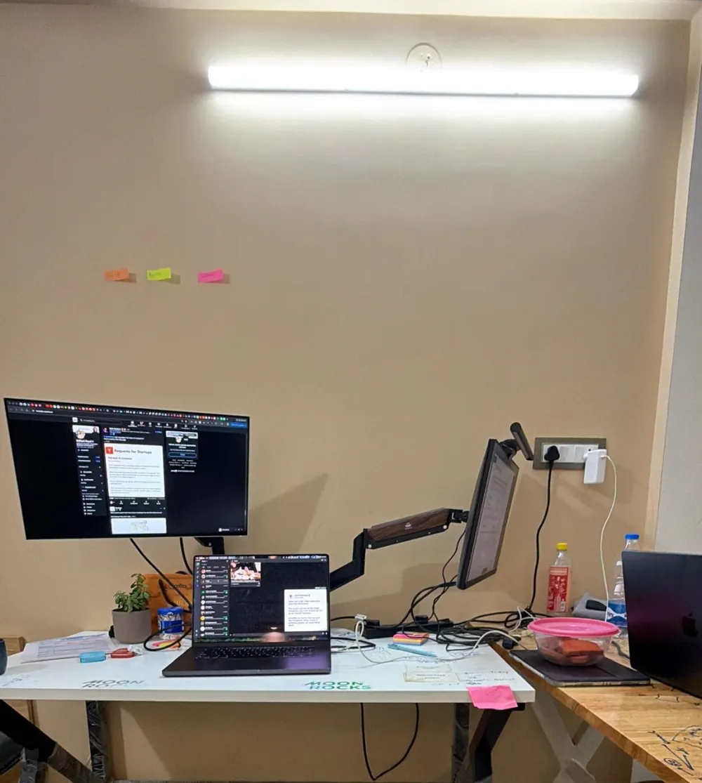 A desk with a laptop and two monitors on arms, three sticky notes on the wall above